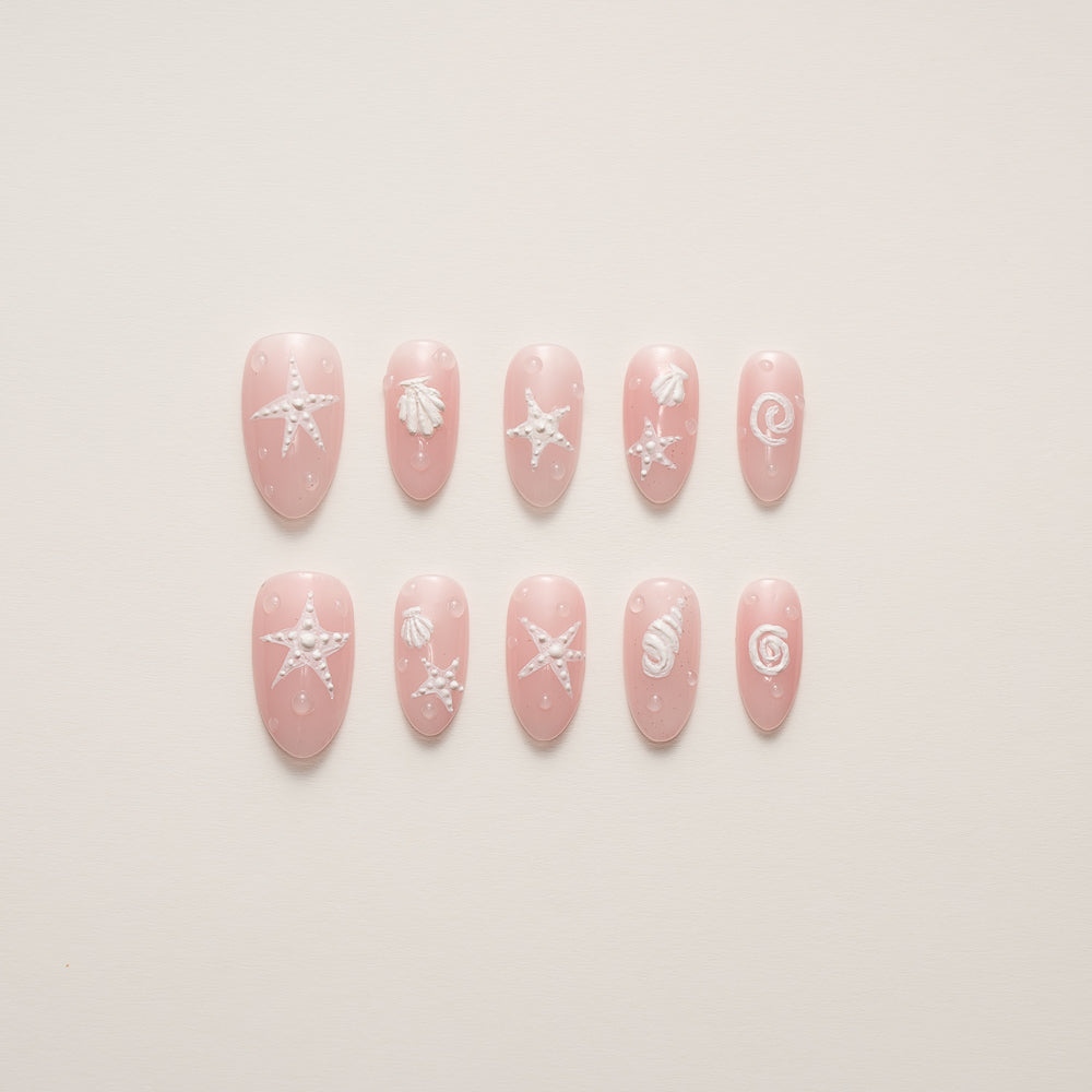 Beachy Pink Press-On Nails with Starfish & Seashell Design resting on a sandy beach backdrop showcasing the coastal manicure set's romantic pink base and marine-inspired charms.