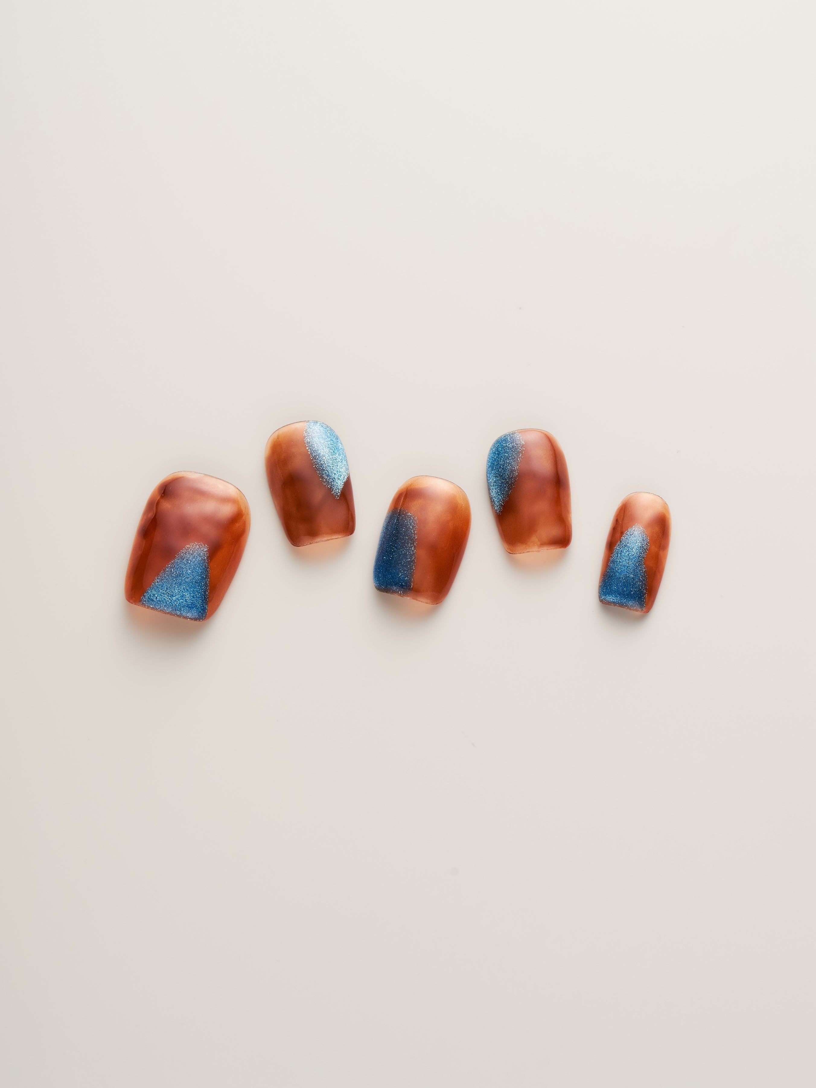 Close-up view of Amber Glow Squoval Press-On Nails in various sizes featuring a blend of amber colors and blue accents.